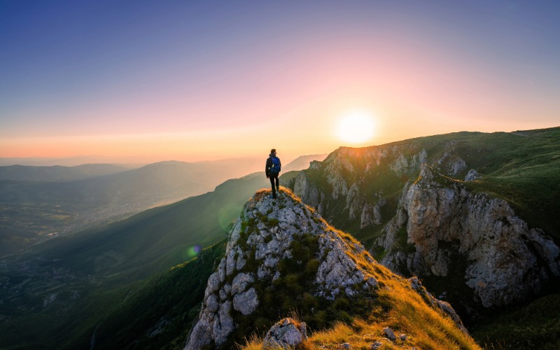 Person standing on top of a mountain overlooking the sunset.